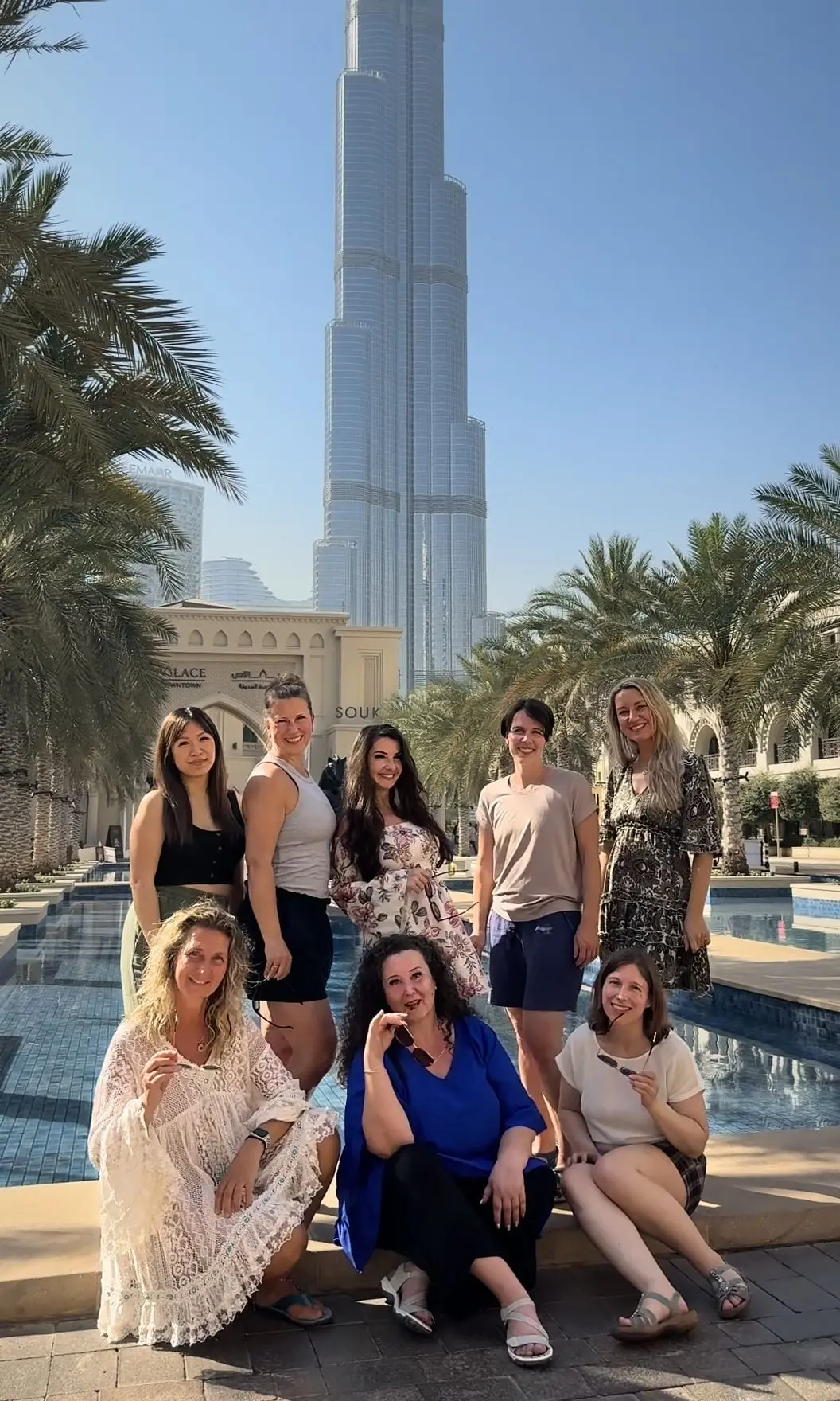 Phoenix in Dubai 2025: acht Frauen an einem Wasserbecken zwischen Palmen, fünf stehen, drei sitzen vorne auf dem Beckenrand, dahinter das Tor zum Souk und der Burj Khalifa
