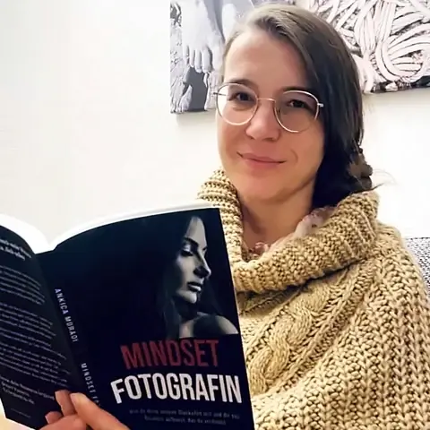 Eva-Maria Preß mit Brille und beigem Strickpullover auf dem Sofa, lächelt und hält das aufgeschlagene Buch „Mindset Fotografin“