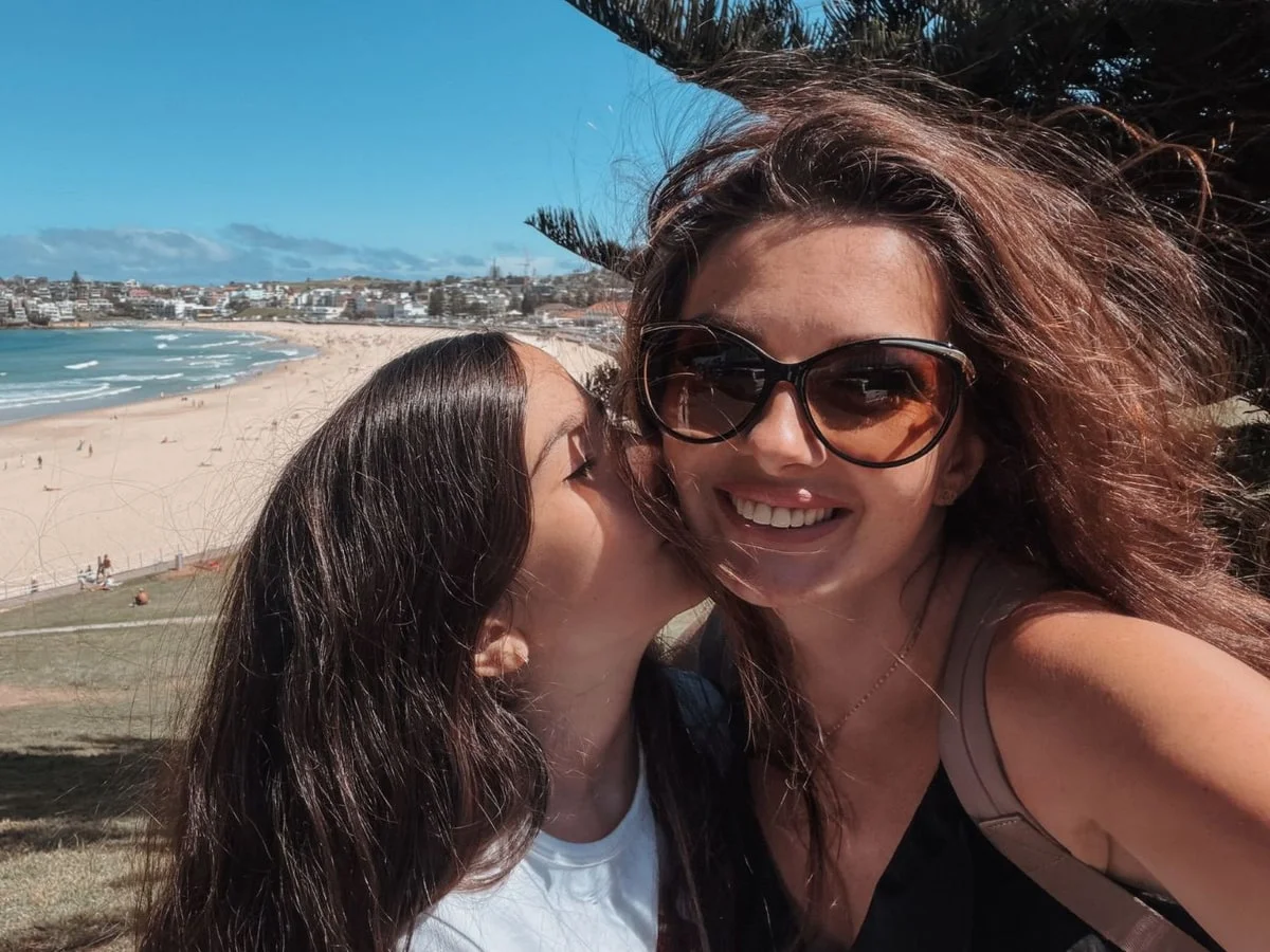 Ankica Moradi mit Sonnenbrille lacht in die Kamera, ihre Tochter küsst sie auf die Wange, dahinter Strand und Meer in Sydney