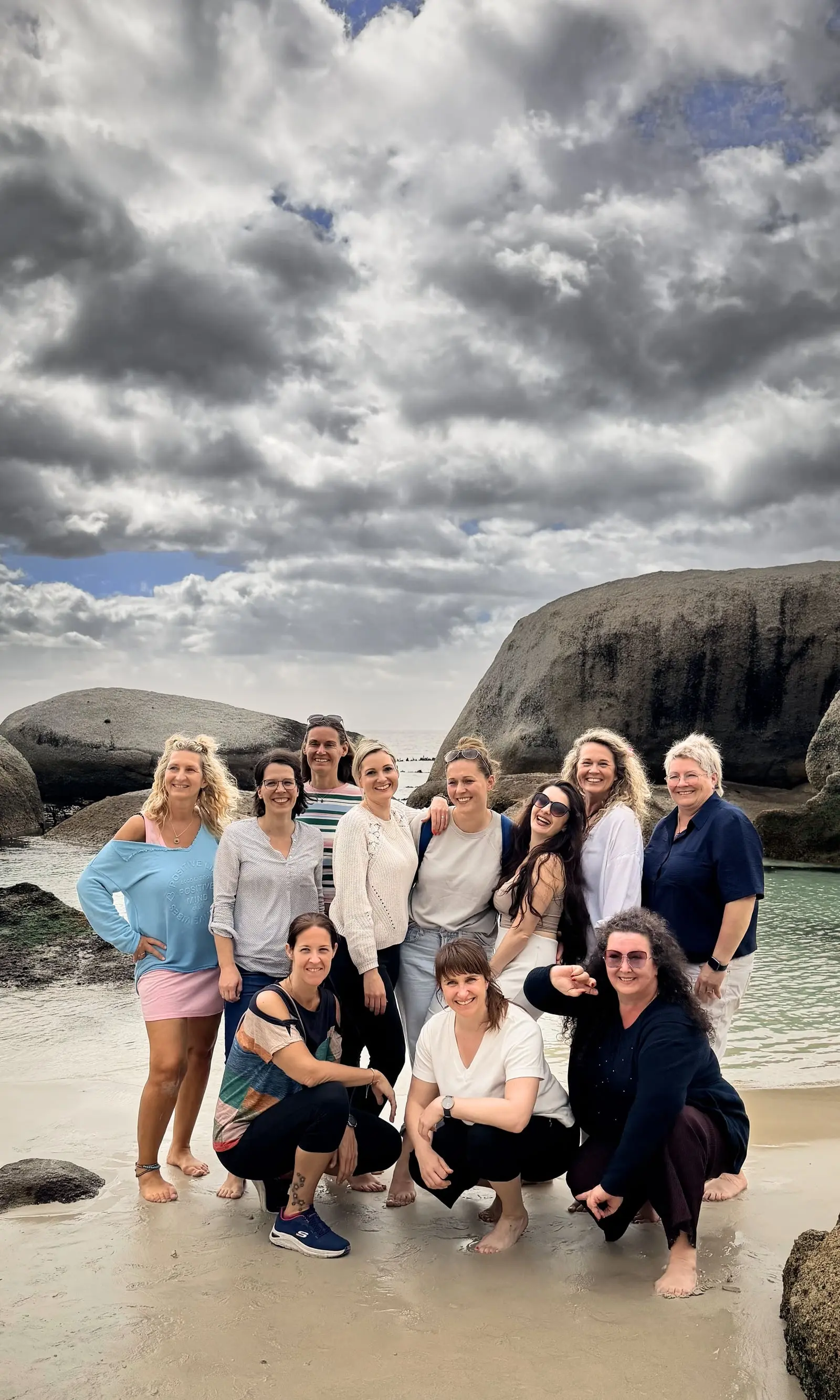 Phoenix in Kapstadt 2026: elf lachende Frauen am Strand zwischen großen Granitfelsen, acht stehen, drei hocken vorne im nassen Sand, dahinter das Meer unter Wolken